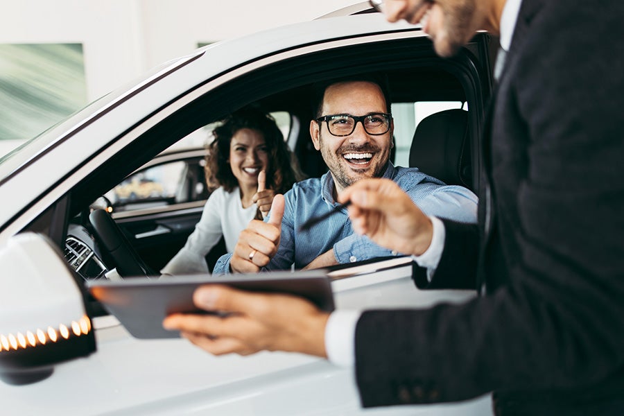 A sales rep in a dark suit bends near the open window and with his Ipad shows a laughing couple seated in the front of the SUV the New GMC vehicle financing options for new buyers